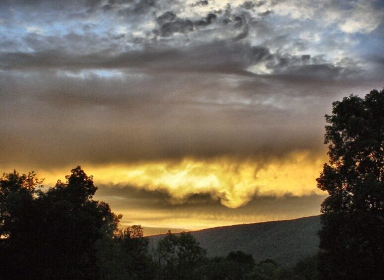 Dramatic sunset sky with dark clouds over silhouetted trees and hills.